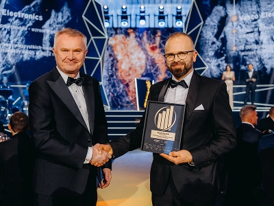 Um plano médio-longo de dois homens vestidos com trajes formais de gala a apertar as mãos. O homem à direita, Maciej Góralski, segura um prémio emoldurado com um emblema dourado, que identifica o prémio como o de «Empreendedor do Ano 2022». No fundo desfocado, um palco apresenta estruturas geométricas abstratas e padrões gráficos dinâmicos em azul e vermelho-alaranjado. Vários convidados estão sentados às mesas no salão de eventos pouco iluminado.