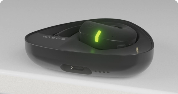 Um auricular Vasco Translator E1 está colocado dentro do seu estojo de carregamento. Os indicadores LED intermitentes mostram o nível atual da bateria do estojo e do auricular. 