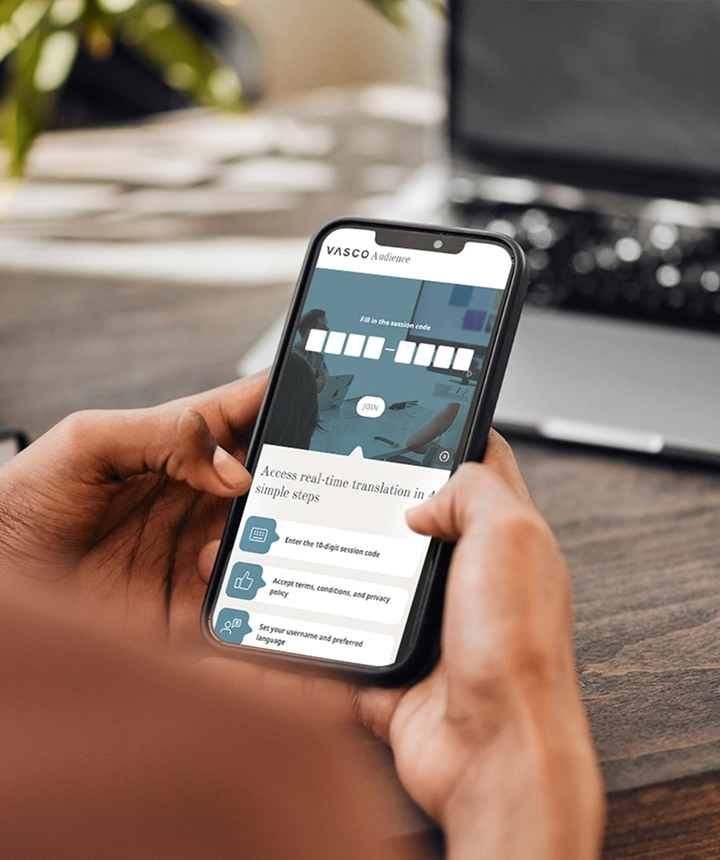 Uma imagem tirada por cima do ombro de uma pessoa a segurar um smartphone que exibe a interface web do Vasco Audience. O ecrã mostra um campo de introdução de dados intitulado «Introduza o código da sessão», com um botão branco «JUNTAR-SE», acima de uma lista de instruções que diz «Aceda à tradução em tempo real em 4 passos simples». O fundo mostra uma secretária de madeira desfocada e um computador portátil.