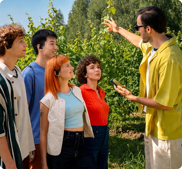 Um grupo de cinco pessoas ao ar livre, num vinhedo banhado pelo sol. À direita, um homem com uma camisa amarela e óculos de sol segura um dispositivo portátil Translator Vasco Q1 e aponta para as videiras verdes. À esquerda, quatro pessoas ouvem atentamente, cada uma com um Translator Vasco E1 preto, demonstrando a funcionalidade de conversação em grupo em tempo real. 