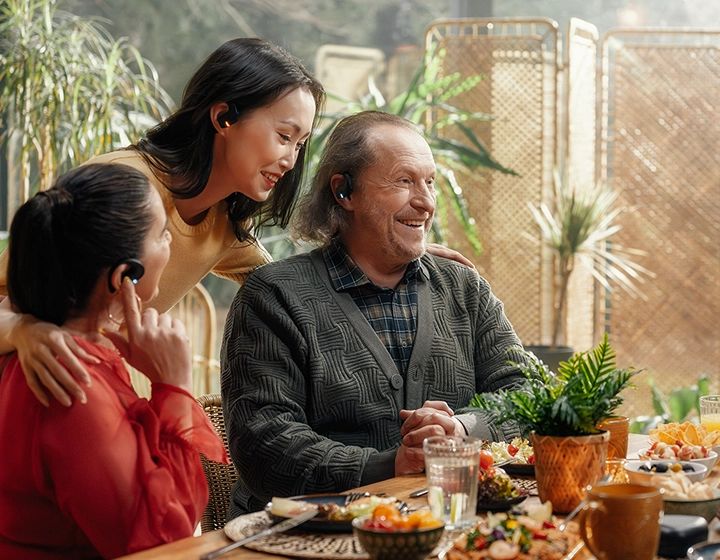 Uma comovente reunião familiar multigeracional à volta de uma mesa de jantar repleta de comida. Um senhor mais velho, sorridente, com um casaco de malha cinzento, e duas mulheres conversam alegremente numa sala iluminada pelo sol e repleta de plantas. Os três têm auscultadores Vasco Translation E1 pretos. 