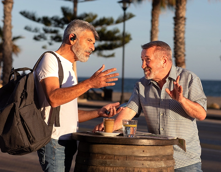 Dois homens estão ao lado de uma mesa ao ar livre a conversar. Um deles tem uma mochila às costas e ambos usam os auriculares Vasco Translator E1. 