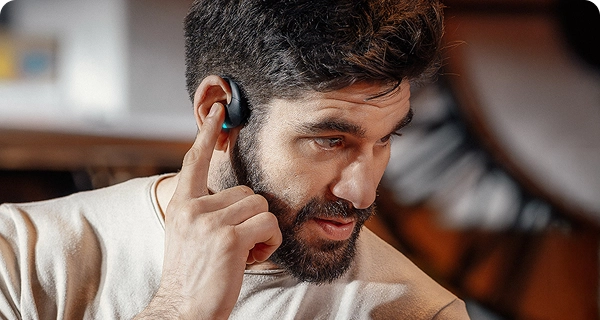 Um grande plano de um homem barbudo a tocar com o dedo indicador num auricular preto de tradução da Vasco, demonstrando os seus controlos táteis intuitivos. Uma pequena luz LED azul brilhante está acesa no dispositivo. 