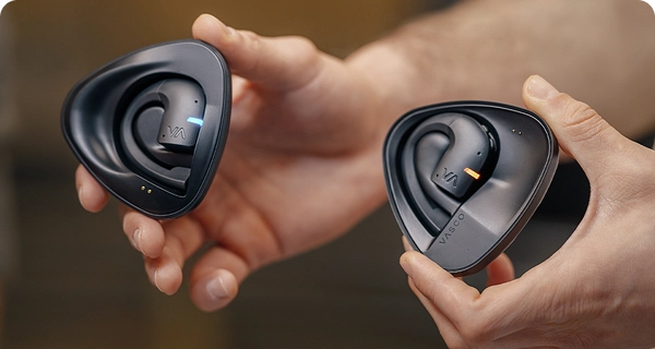 Um grande plano das mãos a segurar dois estojos de carregamento pretos distintos com os auriculares Vasco Translation. A imagem mostra como distinguir os dois, apresentando um auricular com uma luz LED azul e o outro com uma luz LED laranja, indicando os diferentes altifalantes. 