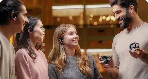 Um grupo de quatro pessoas de diferentes origens apresenta o design universal dos auriculares Vasco Translation E1 numa sala com iluminação acolhedora. Três pessoas têm os auriculares pretos colocados, demonstrando o ajuste confortável para diferentes formatos de orelha, enquanto um homem barbudo e sorridente, à direita, segura dois estojos de carregamento. 