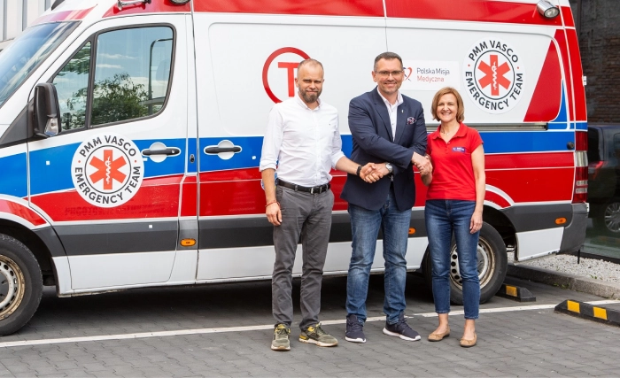 Três pessoas estão sorrindo em frente a uma ambulância branca, vermelha e azul estacionada numa rua pavimentada ao lado de um edifício. Dois homens, um de fato e outro de camisa branca, apertam as mãos, acompanhados por uma mulher de camisa vermelha.