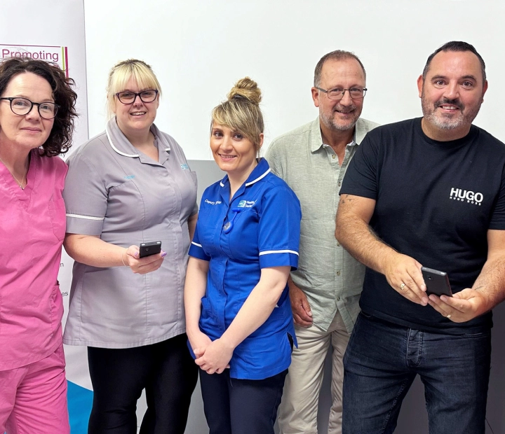 Cinco pessoas sorridentes estão juntas em trajes de saúde, com duas delas segurando um dispositivo tradutor.