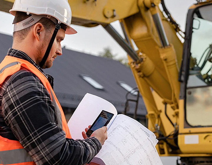 Um homem com capacete e roupa de trabalho a tirar uma fotografia das plantas de construção utilizando o Vasco Translator V4, com uma escavadora visível ao fundo.