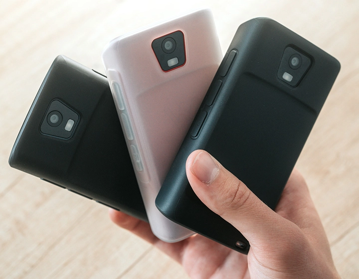 Pessoa segurando três dispositivos Vasco Translator Q1 em capas de silicone, apresentando três cores diferentes de capa: Preto Liso, Caramelo Escuro e Branco Leitoso.