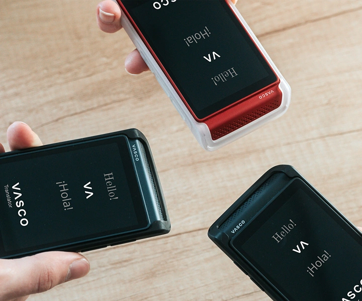 Três pessoas com dispositivos Vasco Translator Q1 juntam-se; os ecrãs mostram a tela inicial com o logótipo da Vasco e as palavras «Hello!» e «¡Hola!». Os dispositivos são apresentados em capas de silicone em três cores: Preto Liso, Caramelo Escuro e Branco Leitoso.