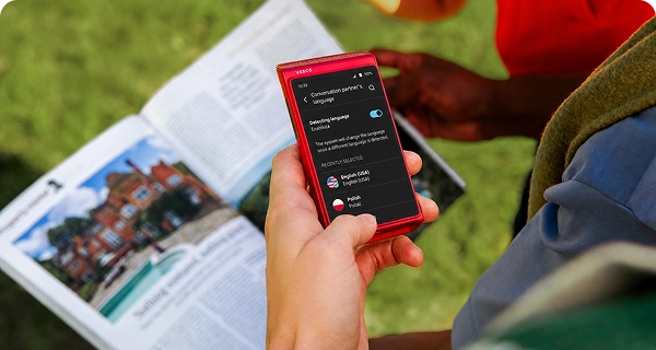 Uma pessoa utiliza um Vasco Translator Q1 vermelho para tirar uma fotografia de um jornal. O ecrã apresenta o menu de seleção de idiomas para traduzir o texto detetado automaticamente no jornal.