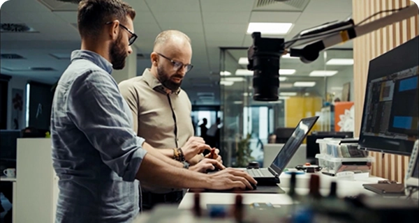 Dois homens com barba e óculos trabalham juntos numa secretária alta, num escritório ou laboratório moderno. Um deles, com uma camisa azul-clara, está a usar um computador portátil, enquanto o outro, com uma camisa bege, está ao seu lado, apontando para um pequeno componente eletrónico que tem na mão. A secretária está equipada com um monitor de grandes dimensões, várias ferramentas eletrónicas e uma câmara montada num braço ajustável. O fundo mostra um espaço de trabalho luminoso e em plano aberto, com divisórias de vidro. 